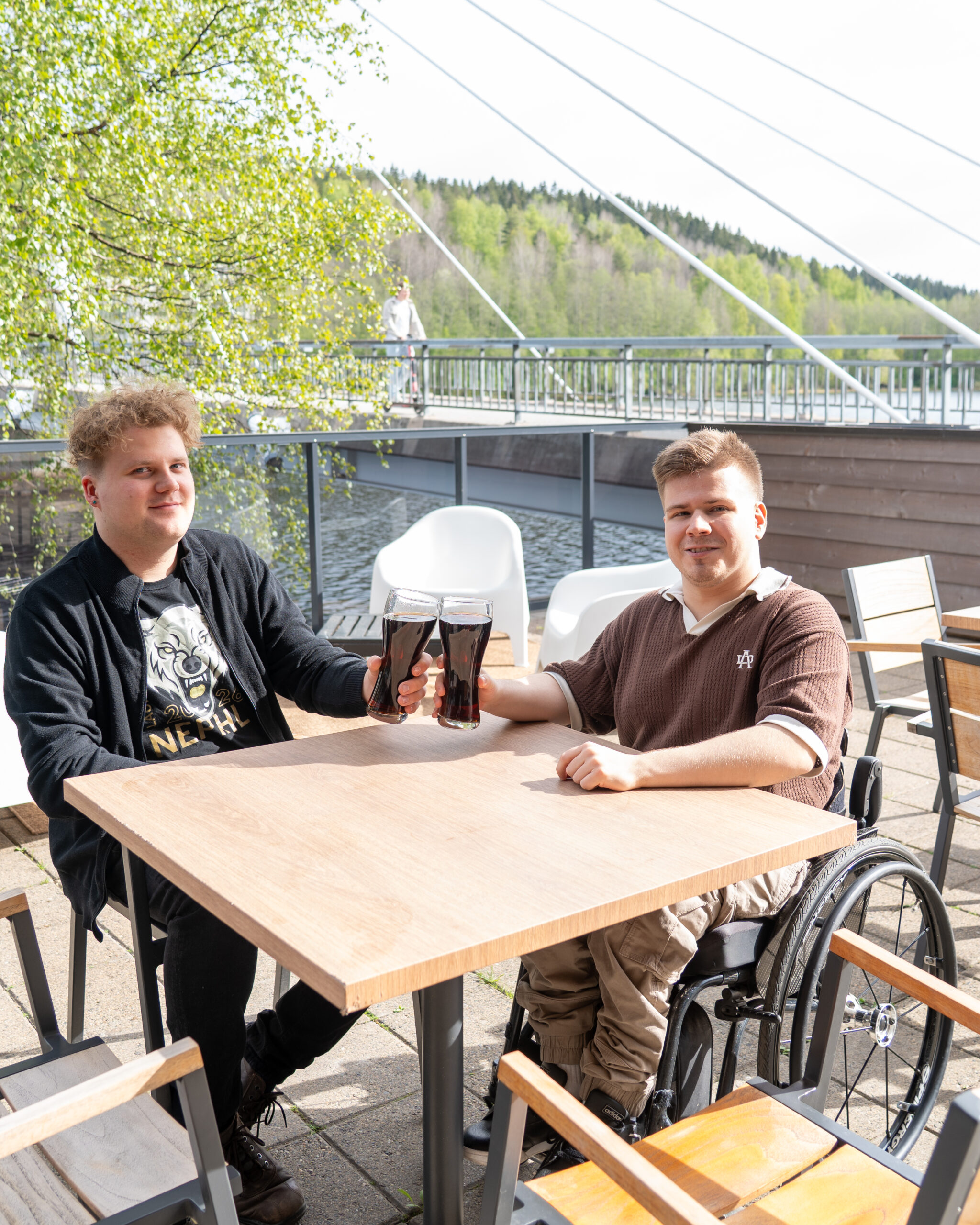 Kaksi miestä oluiden kanssa esteettömällä terassilla, järven rantamaisemissa. Toinen asiakkaista on pyörätuolissa.