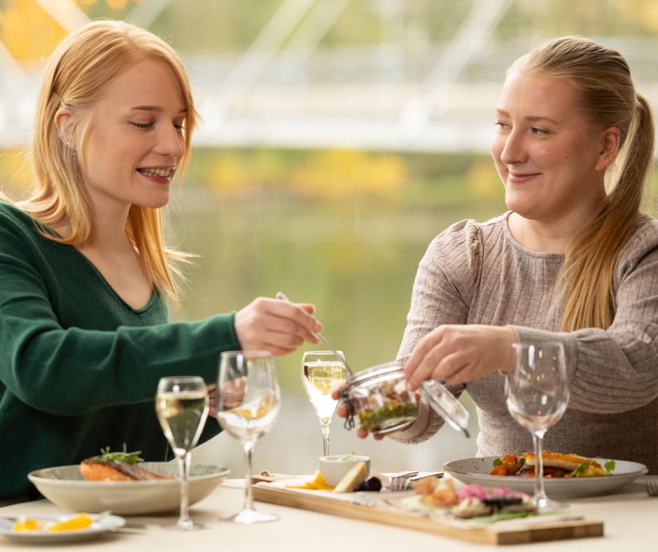 Kaksi iloista naista ruokailee ravintolassa järvimaisemissa